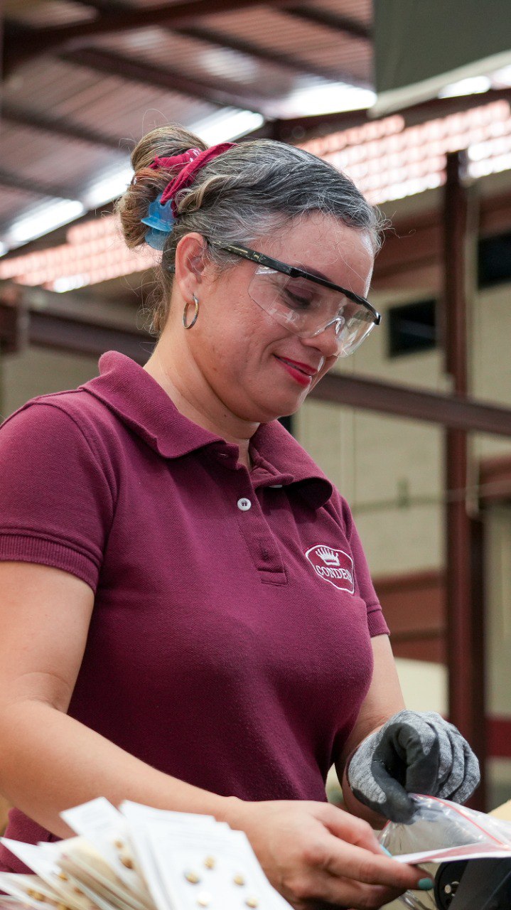 Operaria con lentes de seguridad y uniforme granate sosteniendo hojas de etiquetas en una planta de producción