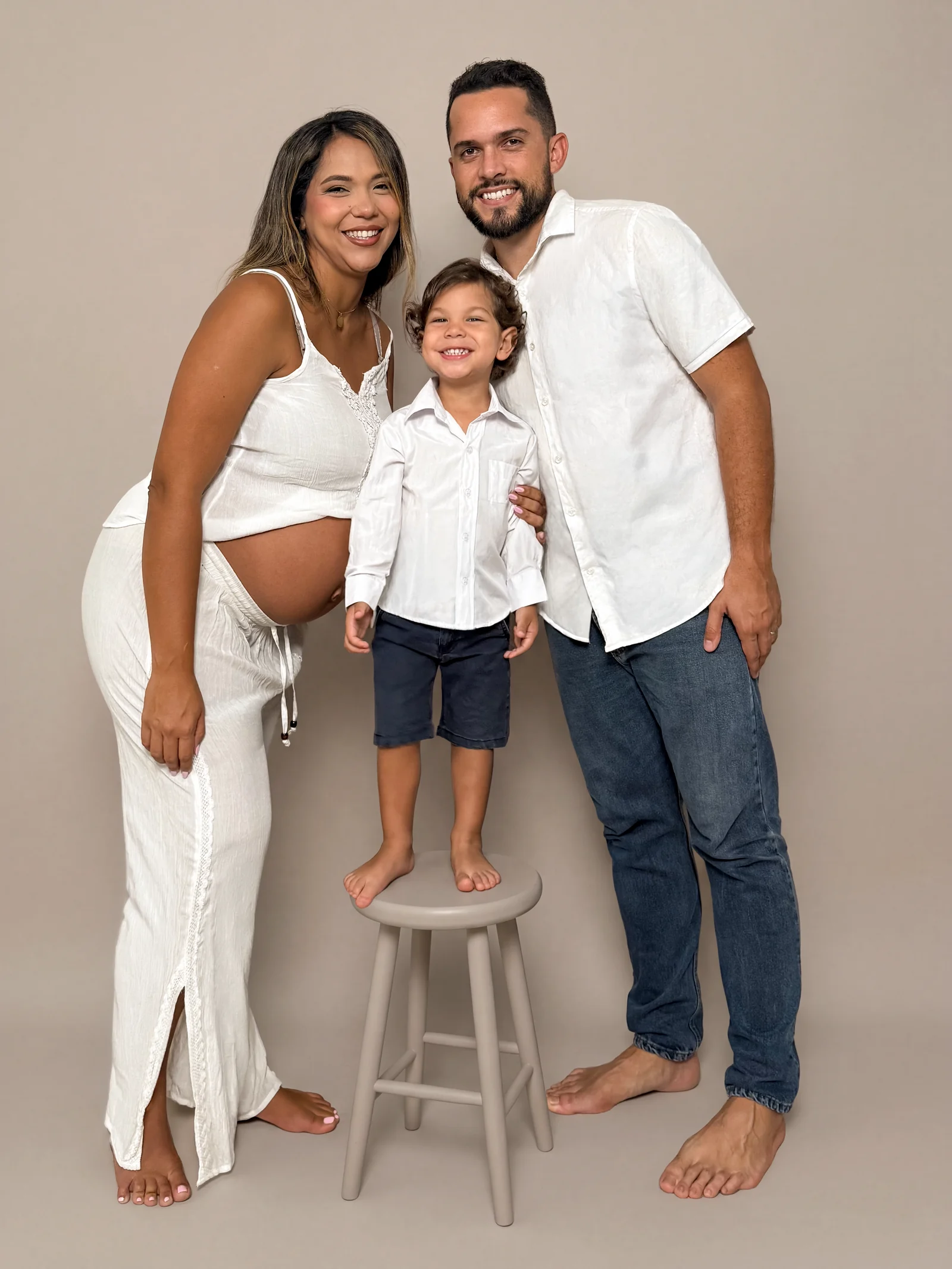 Pareja con su hijo pequeño de pie en estudio, ella embarazada, con atuendo claro sobre fondo beige