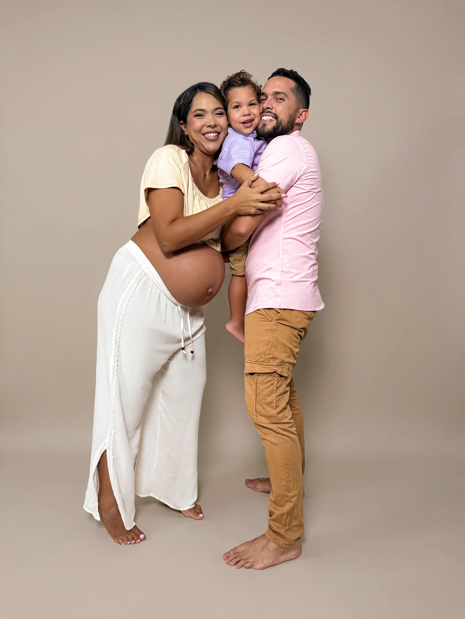 Mujer embarazada y su pareja abrazados, con su hijo pequeño en brazos de él, sobre fondo beige