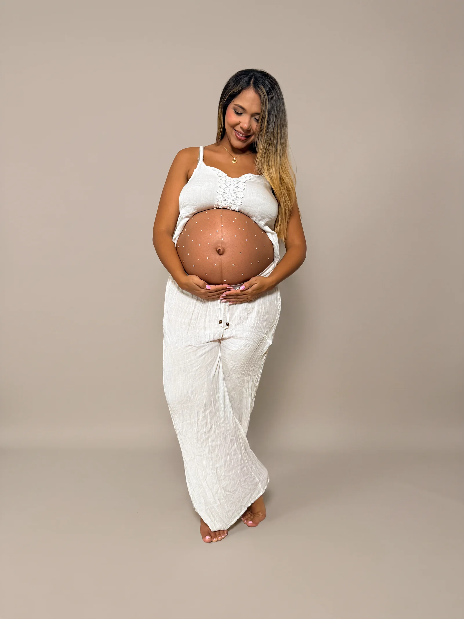 Mujer embarazada de pie con las manos sobre el vientre, con ropa blanca sobre fondo beige